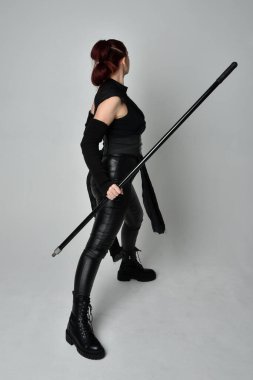 Siyah fütüristik deri kostüm giyen ve elinde mızrak tutan kızıl saçlı güzel bir kadın modelin tam boy portresi. Dinamik duruş pozisyonu ters, beyaz stüdyo arka planı.