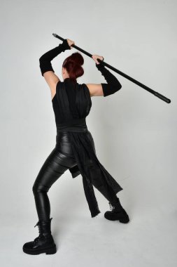 Siyah fütüristik deri kostüm giyen ve elinde mızrak tutan kızıl saçlı güzel bir kadın modelin tam boy portresi. Dinamik duruş pozisyonu ters, beyaz stüdyo arka planı.