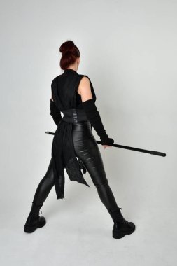 Siyah fütüristik deri kostüm giyen ve elinde mızrak tutan kızıl saçlı güzel bir kadın modelin tam boy portresi. Dinamik duruş pozisyonu ters, beyaz stüdyo arka planı.
