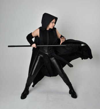 Siyahi fütüristik deri kostüm giyen, elinde mızrak tutan güzel kızıl saçlı kadın modelin tam boy portresi. Beyaz stüdyo arka planında dinamik duruş.