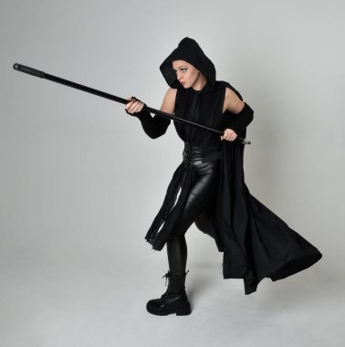Siyahi fütüristik deri kostüm giyen, elinde mızrak tutan güzel kızıl saçlı kadın modelin tam boy portresi. Beyaz stüdyo arka planında dinamik duruş.