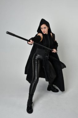 Siyahi fütüristik deri kostüm giyen, elinde mızrak tutan güzel kızıl saçlı kadın modelin tam boy portresi. Beyaz stüdyo arka planında dinamik duruş.
