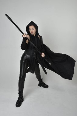 Siyahi fütüristik deri kostüm giyen, elinde mızrak tutan güzel kızıl saçlı kadın modelin tam boy portresi. Beyaz stüdyo arka planında dinamik duruş.