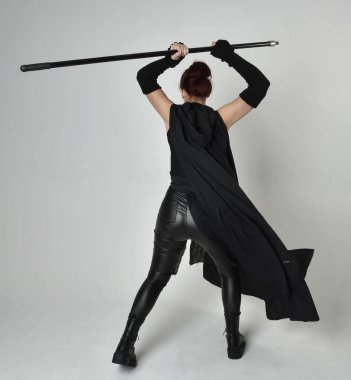 Siyahi fütüristik deri kostüm giyen, elinde mızrak tutan güzel kızıl saçlı kadın modelin tam boy portresi. Beyaz stüdyo arka planında dinamik duruş.