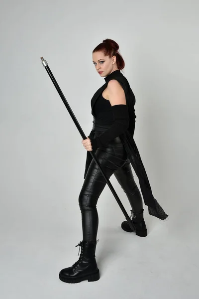 全长肖像漂亮红头发的女模特身穿黑色未来科幻皮衣 手持长矛武器 动态站立姿势 面朝外 背向后 白色工作室背景 — 图库照片