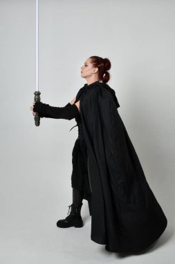 Siyah fütüristik deri pelerin giymiş, elinde ışın kılıcı olan güzel kızıl saçlı kadın modelin tam boy portresi. Beyaz stüdyo arka planında dinamik duruş.