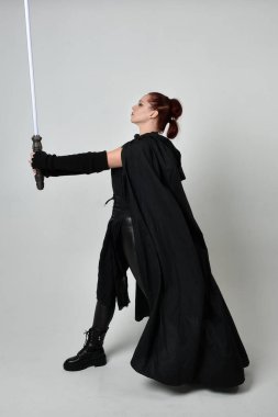 Siyah fütüristik deri pelerin giymiş, elinde ışın kılıcı olan güzel kızıl saçlı kadın modelin tam boy portresi. Beyaz stüdyo arka planında dinamik duruş.