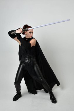 Siyah fütüristik deri pelerin giymiş, elinde ışın kılıcı olan güzel kızıl saçlı kadın modelin tam boy portresi. Beyaz stüdyo arka planında dinamik duruş.