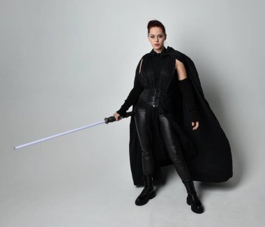 Siyah fütüristik deri pelerin giymiş, elinde ışın kılıcı olan güzel kızıl saçlı kadın modelin tam boy portresi. Beyaz stüdyo arka planında dinamik duruş.