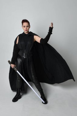 Full length portrait of pretty redhead female model wearing black futuristic scifi leather cloak costume, holding a lightsaber weapon. Dynamic standing pose on white studio background. clipart