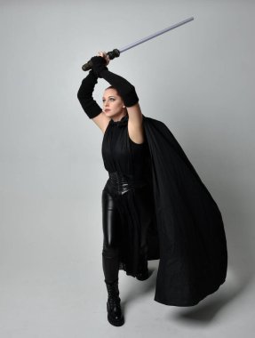 Siyah fütüristik deri pelerin giymiş, elinde ışın kılıcı olan güzel kızıl saçlı kadın modelin tam boy portresi. Beyaz stüdyo arka planında dinamik duruş.