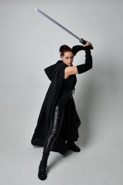 Siyah fütüristik deri pelerin giymiş, elinde ışın kılıcı olan güzel kızıl saçlı kadın modelin tam boy portresi. Beyaz stüdyo arka planında dinamik duruş.