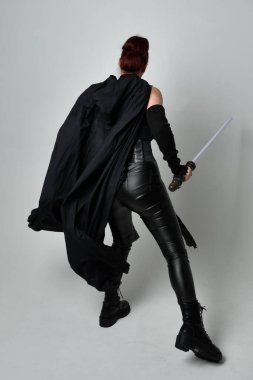 Siyah fütüristik deri pelerin giymiş, elinde ışın kılıcı olan güzel kızıl saçlı kadın modelin tam boy portresi. Beyaz stüdyo arka planında dinamik duruş.