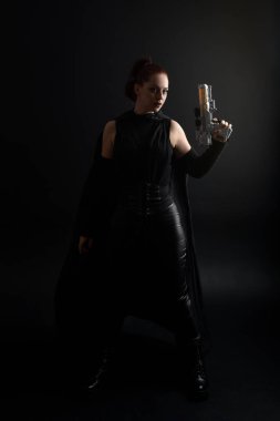 Siyahi fütürist deri pelerin giymiş, elinde silah tutan kızıl saçlı güzel bir kadın modelin tam boy portresi. Stüdyonun arka planında dinamik duruş.