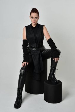 Siyah fütüristik deri pelerin giyen güzel kızıl saçlı kadın modelin tam boy portresi. Dinamik oturma beyaz stüdyo arka planı üzerine diz çökmüş poz.