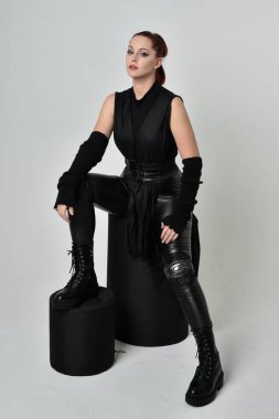 Siyah fütüristik deri pelerin giyen güzel kızıl saçlı kadın modelin tam boy portresi. Dinamik oturma beyaz stüdyo arka planı üzerine diz çökmüş poz.