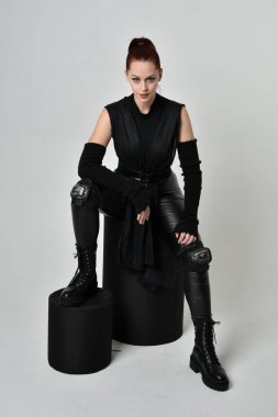 Siyah fütüristik deri pelerin giyen güzel kızıl saçlı kadın modelin tam boy portresi. Dinamik oturma beyaz stüdyo arka planı üzerine diz çökmüş poz.