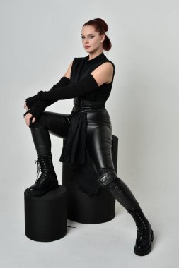 Siyah fütüristik deri pelerin giyen güzel kızıl saçlı kadın modelin tam boy portresi. Dinamik oturma beyaz stüdyo arka planı üzerine diz çökmüş poz.