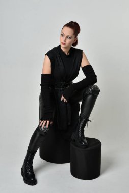 Siyah fütüristik deri pelerin giyen güzel kızıl saçlı kadın modelin tam boy portresi. Dinamik oturma beyaz stüdyo arka planı üzerine diz çökmüş poz.