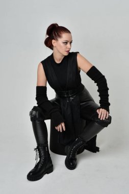 Siyah fütüristik deri pelerin giyen güzel kızıl saçlı kadın modelin tam boy portresi. Dinamik oturma beyaz stüdyo arka planı üzerine diz çökmüş poz.