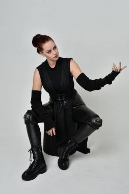 Siyah fütüristik deri pelerin giyen güzel kızıl saçlı kadın modelin tam boy portresi. Dinamik oturma beyaz stüdyo arka planı üzerine diz çökmüş poz.