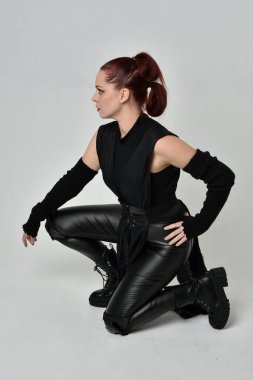 Siyah fütüristik deri pelerin giyen güzel kızıl saçlı kadın modelin tam boy portresi. Dinamik oturma beyaz stüdyo arka planı üzerine diz çökmüş poz.
