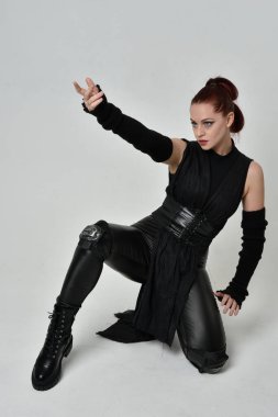 Siyah fütüristik deri pelerin giyen güzel kızıl saçlı kadın modelin tam boy portresi. Dinamik oturma beyaz stüdyo arka planı üzerine diz çökmüş poz.