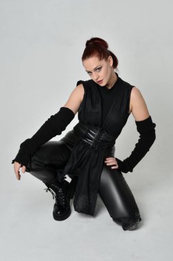 Siyah fütüristik deri pelerin giyen güzel kızıl saçlı kadın modelin tam boy portresi. Dinamik oturma beyaz stüdyo arka planı üzerine diz çökmüş poz.