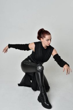 Siyah fütüristik deri pelerin giyen güzel kızıl saçlı kadın modelin tam boy portresi. Dinamik oturma beyaz stüdyo arka planı üzerine diz çökmüş poz.