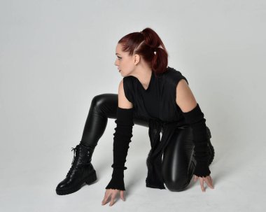Siyah fütüristik deri pelerin giyen güzel kızıl saçlı kadın modelin tam boy portresi. Dinamik oturma beyaz stüdyo arka planı üzerine diz çökmüş poz.