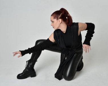 Siyah fütüristik deri pelerin giyen güzel kızıl saçlı kadın modelin tam boy portresi. Dinamik oturma beyaz stüdyo arka planı üzerine diz çökmüş poz.