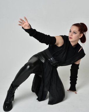 Siyah fütüristik deri pelerin giyen güzel kızıl saçlı kadın modelin tam boy portresi. Dinamik oturma beyaz stüdyo arka planı üzerine diz çökmüş poz.