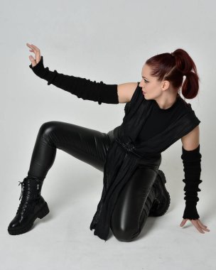 Siyah fütüristik deri pelerin giyen güzel kızıl saçlı kadın modelin tam boy portresi. Dinamik oturma beyaz stüdyo arka planı üzerine diz çökmüş poz.
