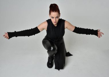 Siyah fütüristik deri pelerin giyen güzel kızıl saçlı kadın modelin tam boy portresi. Dinamik oturma beyaz stüdyo arka planı üzerine diz çökmüş poz.