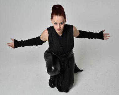 Siyah fütüristik deri pelerin giyen güzel kızıl saçlı kadın modelin tam boy portresi. Dinamik oturma beyaz stüdyo arka planı üzerine diz çökmüş poz.