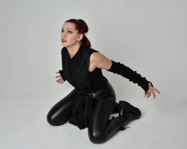 Siyah fütüristik deri pelerin giyen güzel kızıl saçlı kadın modelin tam boy portresi. Dinamik oturma beyaz stüdyo arka planı üzerine diz çökmüş poz.