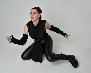 Siyah fütüristik deri pelerin giyen güzel kızıl saçlı kadın modelin tam boy portresi. Dinamik oturma beyaz stüdyo arka planı üzerine diz çökmüş poz.