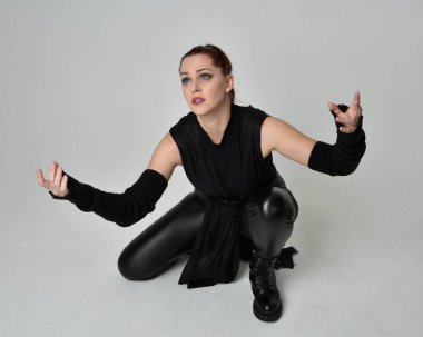 Siyah fütüristik deri pelerin giyen güzel kızıl saçlı kadın modelin tam boy portresi. Dinamik oturma beyaz stüdyo arka planı üzerine diz çökmüş poz.