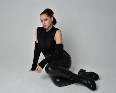 Siyah fütüristik deri pelerin giyen güzel kızıl saçlı kadın modelin tam boy portresi. Dinamik oturma beyaz stüdyo arka planı üzerine diz çökmüş poz.