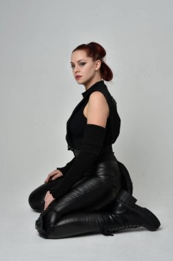 Siyah fütüristik deri pelerin giyen güzel kızıl saçlı kadın modelin tam boy portresi. Dinamik oturma beyaz stüdyo arka planı üzerine diz çökmüş poz.