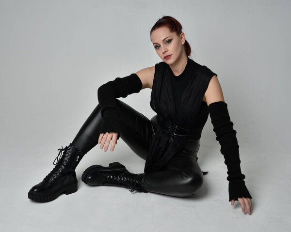 Полнометражный портрет рыжеволосой красавицы-модели в черном футуристическом кожаном плаще. Динамичная сидящая на коленях поза на белом фоне студии.