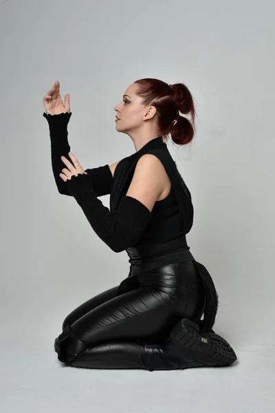 黒未来的なScifi革のマントの衣装を身に着けているかなり赤毛の女性モデルの完全な長さの肖像画 ダイナミック座位膝ポーズ上のホワイトスタジオの背景 — ストック写真