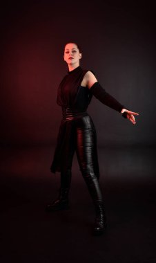 Siyah fütüristik deri pelerin giyen güzel kızıl saçlı kadın modelin tam boy portresi. Karanlık stüdyo arka planında, karamsar ışıklandırmalı duruş..