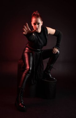 Siyah fütüristik deri pelerin giyen güzel kızıl saçlı kadın modelin tam boy portresi. Karanlık stüdyo arka planında oturan karamsar ışıklandırmalı..