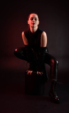 Siyah fütüristik deri pelerin giyen güzel kızıl saçlı kadın modelin tam boy portresi. Karanlık stüdyo arka planında oturan karamsar ışıklandırmalı..