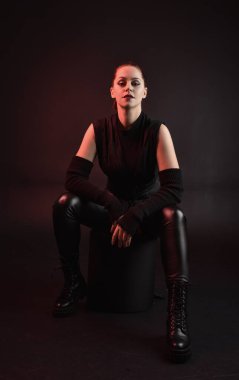 Siyah fütüristik deri pelerin giyen güzel kızıl saçlı kadın modelin tam boy portresi. Karanlık stüdyo arka planında oturan karamsar ışıklandırmalı..