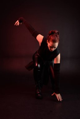 Siyah fütüristik deri pelerin giyen güzel kızıl saçlı kadın modelin tam boy portresi. Karanlık stüdyo arka planında oturan karamsar ışıklandırmalı..