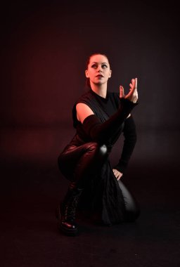 Siyah fütüristik deri pelerin giyen güzel kızıl saçlı kadın modelin tam boy portresi. Karanlık stüdyo arka planında oturan karamsar ışıklandırmalı..