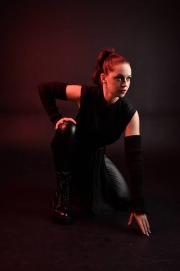 Siyah fütüristik deri pelerin giyen güzel kızıl saçlı kadın modelin tam boy portresi. Karanlık stüdyo arka planında oturan karamsar ışıklandırmalı..