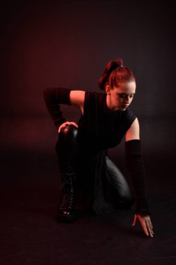 Siyah fütüristik deri pelerin giyen güzel kızıl saçlı kadın modelin tam boy portresi. Karanlık stüdyo arka planında oturan karamsar ışıklandırmalı..